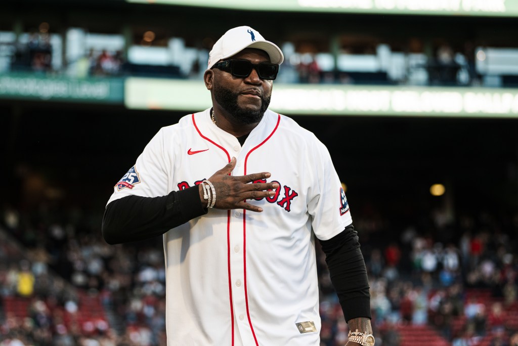 David Ortiz in a white Boston Red Sox jersey, sunglasses, and a cap, with his hand over his heart during a pregame ceremony.