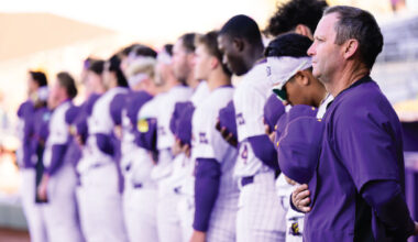 Lineup of athletes in purple and white uniforms standing in a row as a coach watches from the foreground at a stadium.