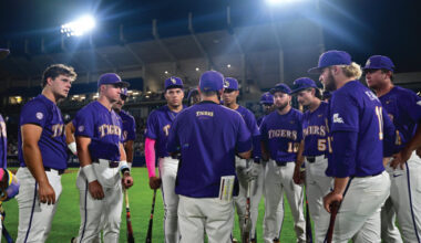 Purple-uniform baseball players form a circle around their coach on the field at night under bright stadium lights, listening intently during a team huddle.