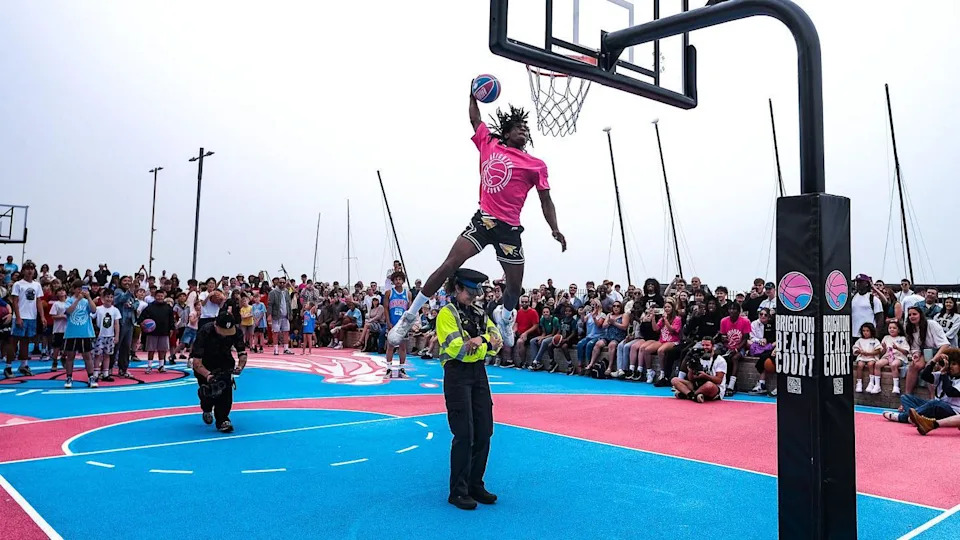 A person in a pink shirt jumping over a female police officer and dunking a basketball on a blue and pink court.