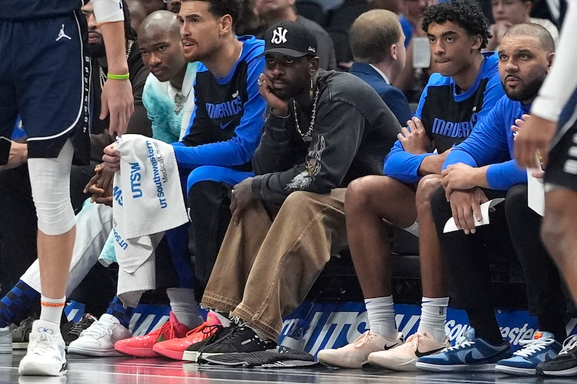 An injured Dallas Mavericks guard Kyrie Irving, center, sits on the bench between teammates...