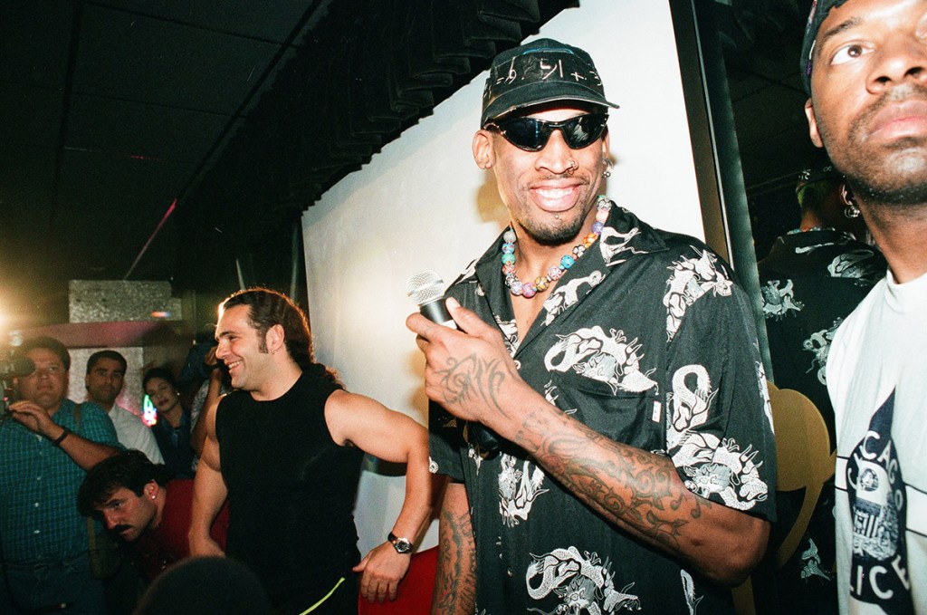 Dennis Rodman onstage at illusions nightclub wearing a baseball cap and sunglasses. he is in the middle of the frame and there is a person to his right and left