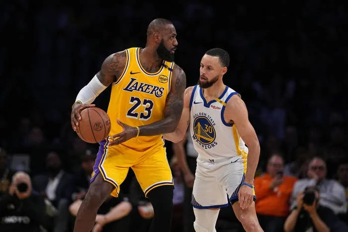 Two basketball players in action. The player on the left wears a Lakers jersey and holds the ball. The player on the right defends in a Warriors uniform