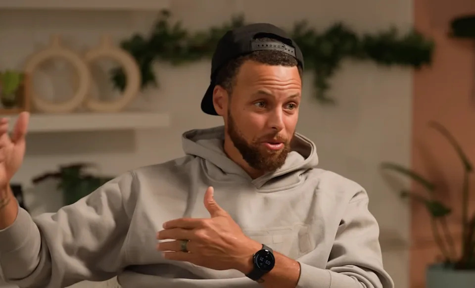 A person wearing a hoodie and backwards cap gestures while talking indoors