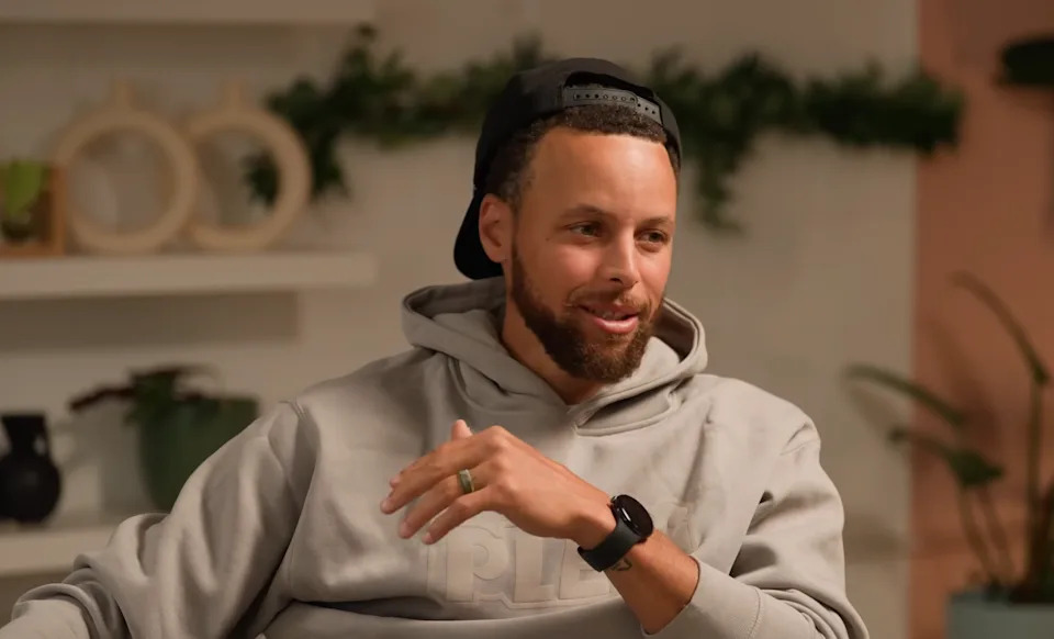 Person in casual hoodie and backwards cap, sitting and smiling in a relaxed setting with plants in the background