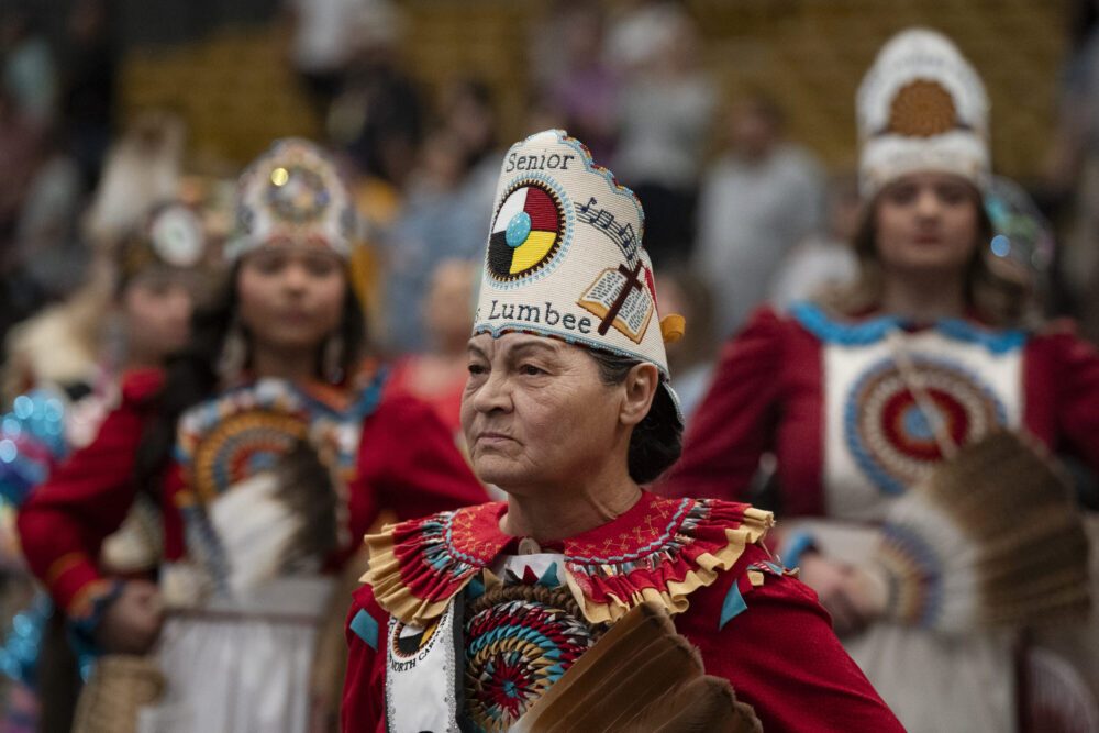 A Lumbee elder poses in regali with a red shirt and white cap with the medicine wheel embroidered on it.