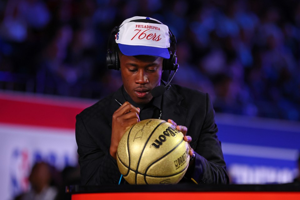A man in a Philadelphia 76ers hat signs a gold basketball.