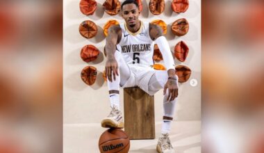 A basketball player in a white New Orleans uniform poses seated on a wooden block, with one hand on his knee and the other resting on a basketball. Behind him, several deflated basketballs are mounted on the wall.