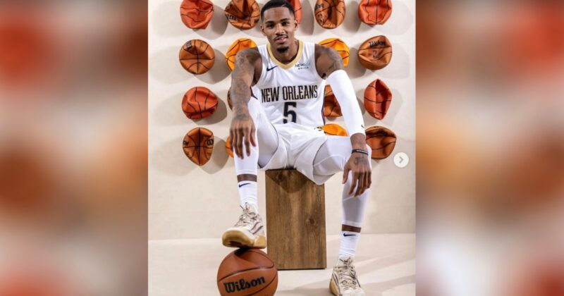 A basketball player in a white New Orleans uniform poses seated on a wooden block, with one hand on his knee and the other resting on a basketball. Behind him, several deflated basketballs are mounted on the wall.