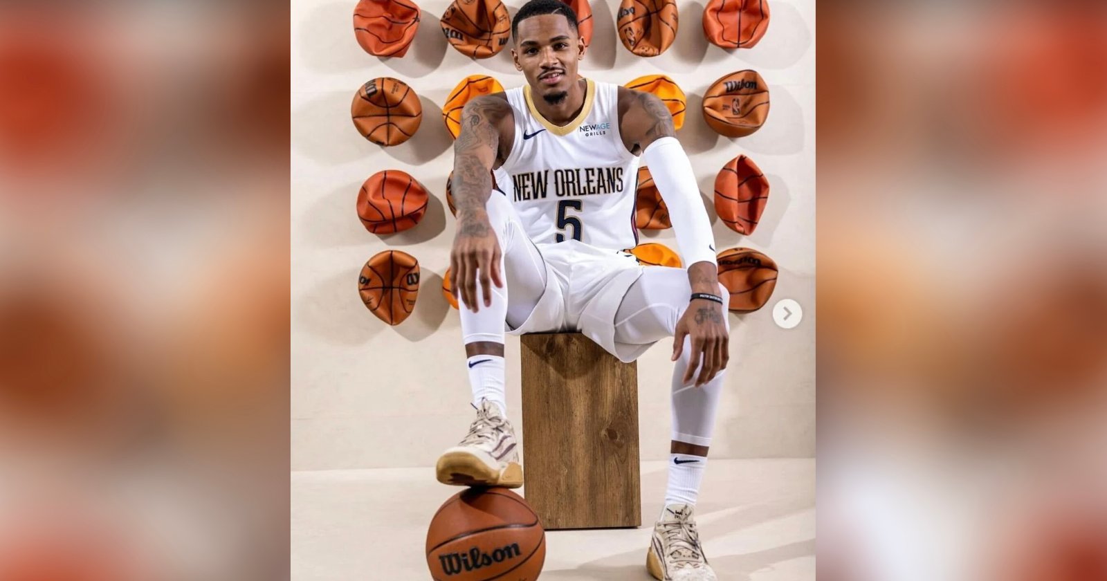 A basketball player in a white New Orleans uniform poses seated on a wooden block, with one hand on his knee and the other resting on a basketball. Behind him, several deflated basketballs are mounted on the wall.
