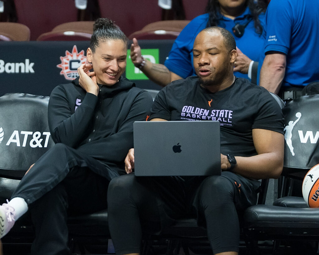 Golden State Valkyries assistant coach Landon Tatum sits on the bench with forward Cecilia Zandalasini before a game. Zandalasini is smiling as they both look at a laptop that Tatum balances on his lap.