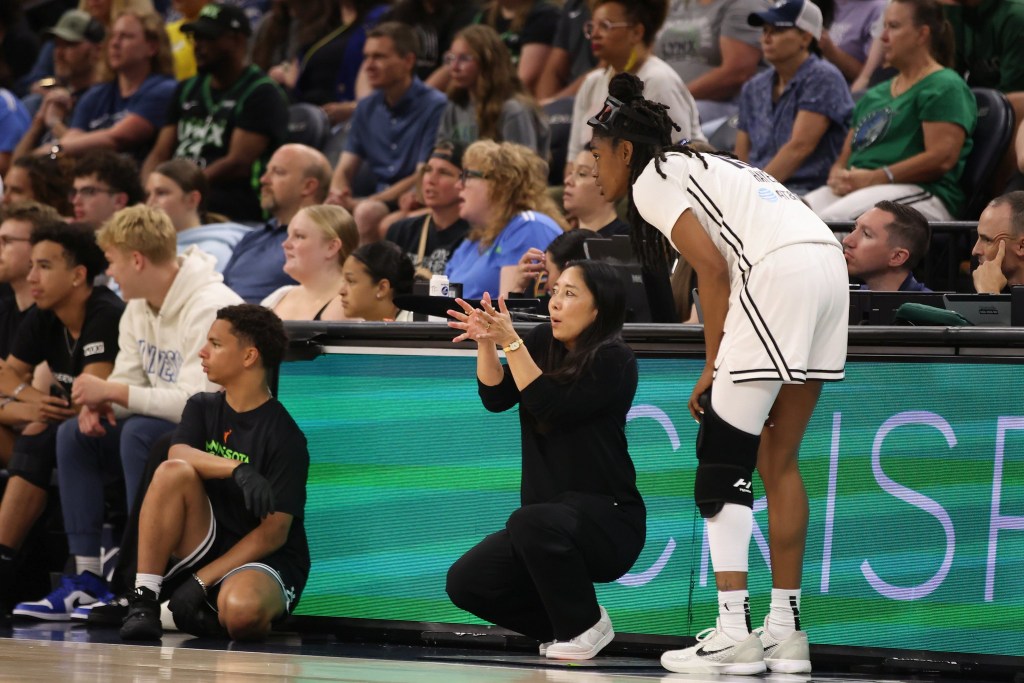 Golden State Valkyries head coach Natalie Nakase squats in front of the scorer's table and talks to guard Tiffany Hayes, who is standing next to her and leaning toward her. Both are looking out in front of them, not at each other, and Nakase has her hands up, just below eye level.