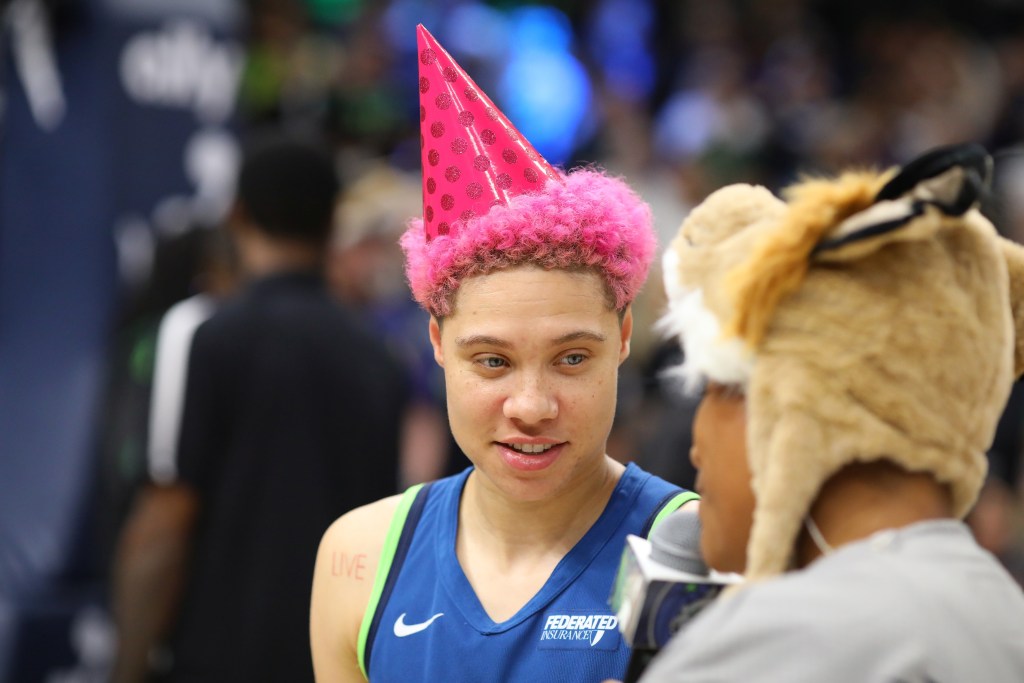 Minnesota Lynx guard Natisha Hiedeman listens to a question from someone holding a Lynx-branded microphone after a game. Hiedeman is still in her blue game jersey but has also added a hot pink polka-dot hat on top of her pink hair.