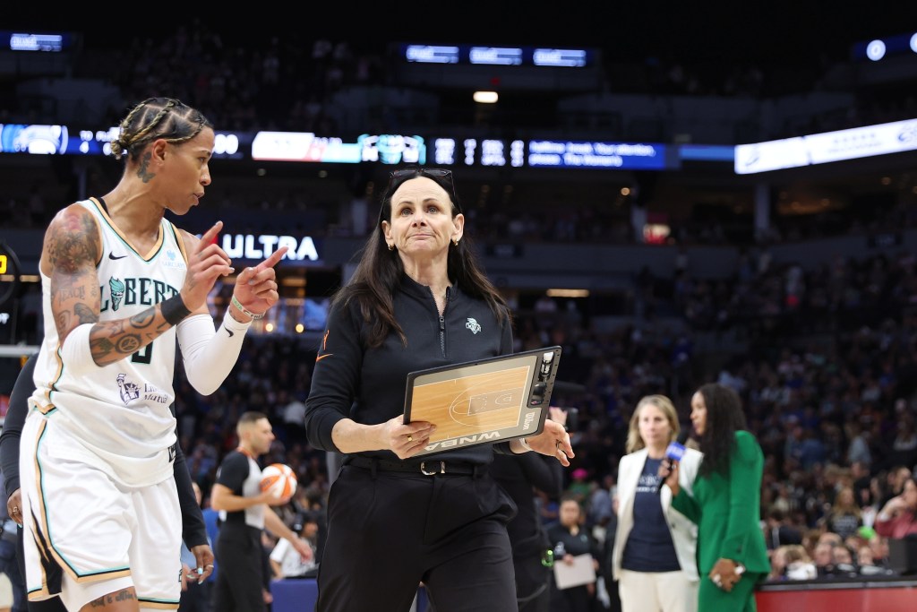 New York Liberty head coach Sandy Brondello holds her coaching board in front of her body while guard Natasha Cloud stands next to her and asks her something.