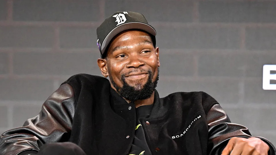 Kevin Durant, wearing a black cap and jacket, smiling during an event.