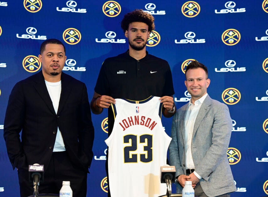 Denver Nuggets basketball player, Cameron Johnson, center, holds his New Jersey during his introductory press conference at Ball Arena in Denver, Colorado, on Friday, July 18, 2025. Jon Wallace, Nuggets' executive vice president of player personnel, left, and Ben Tenzer, right, Nuggets' executive vice president of basketball operations, join Johnson on stage.
