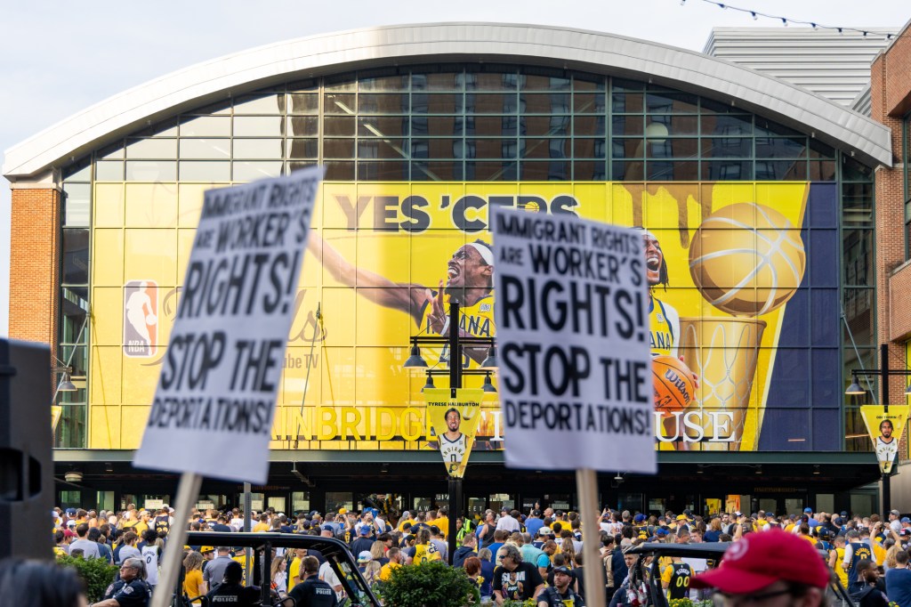 Pacers fans crowd the plaza outside Gainbridge Fieldhouse. in the foreground, protest signs read, "Immigrant rights are worker's rights! Stop the deportations!"