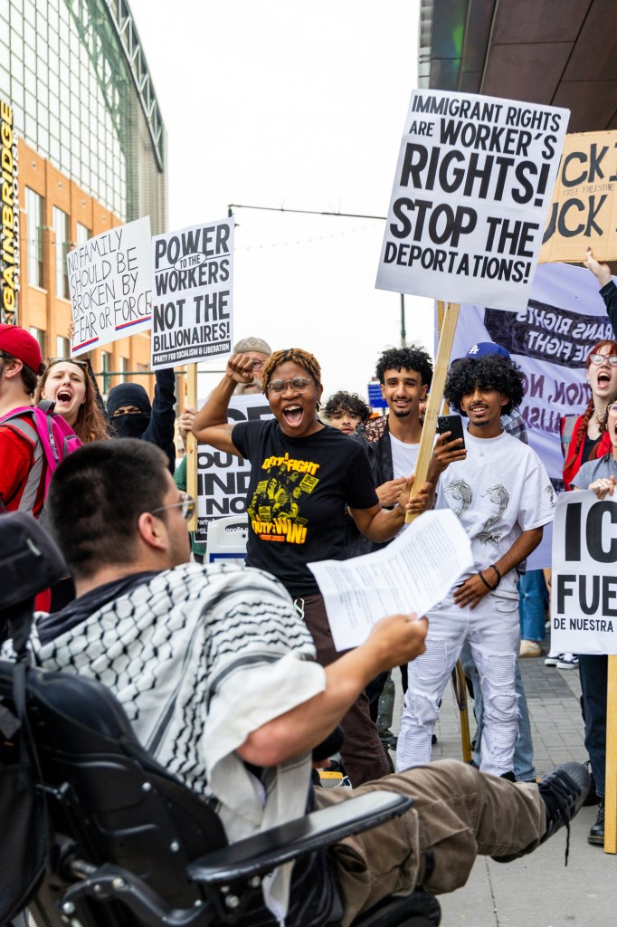 Protesters stand on a city street facing a person in a wheelchair, cheering and holding signs that read things like, "Power to the workers not the billionaires!" "No family should be broken by fear or force."