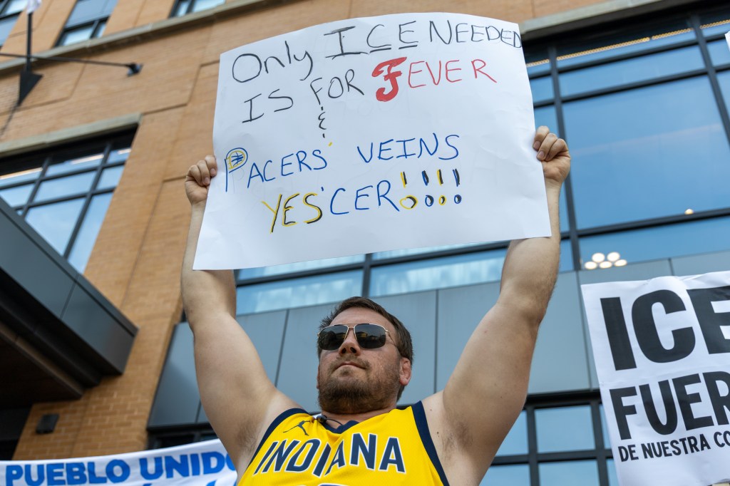 A man wearing an Indiana Pacers jersey and sunglasses holds a poster over his head reading, "Only ICE neede is for Fever and Pacers' veins yes'cer!"