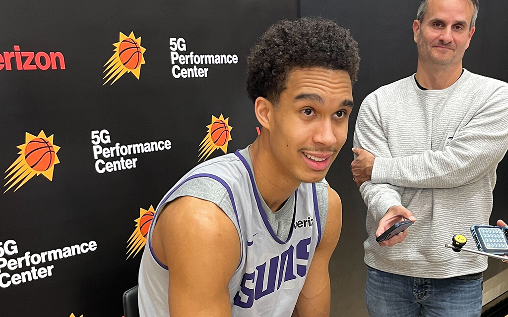 Basketball player in a gray and purple Phoenix Suns jersey smiling in front of a branded backdrop.