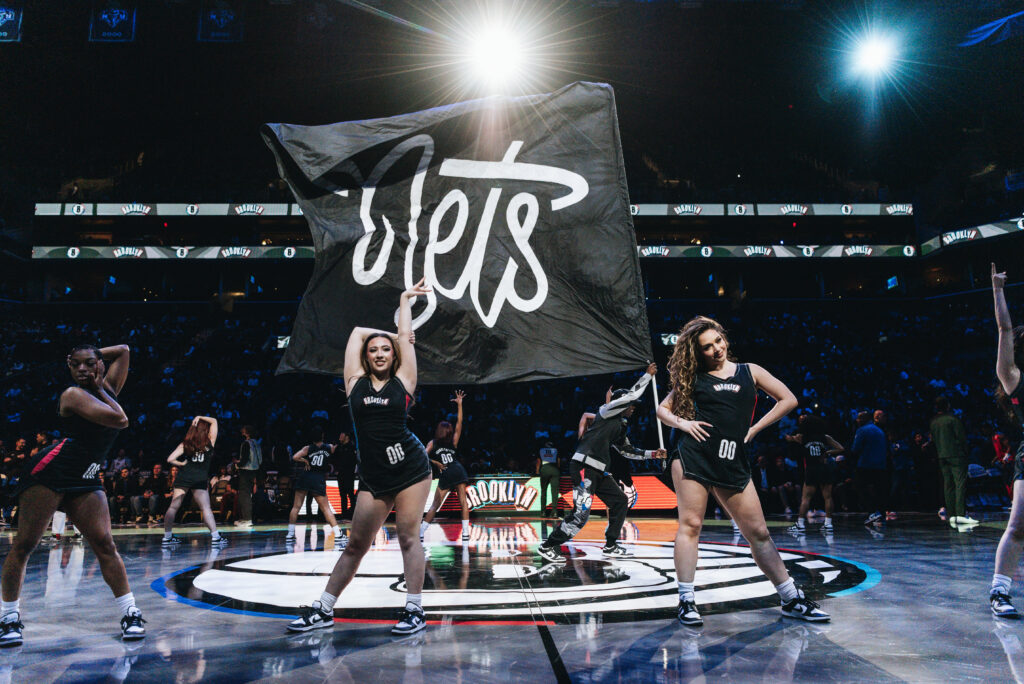 Under brights lights on a basketball court, members of a dance team stand proudly across the court in fierce poses as a man waves a large Nets fan in the middle.
