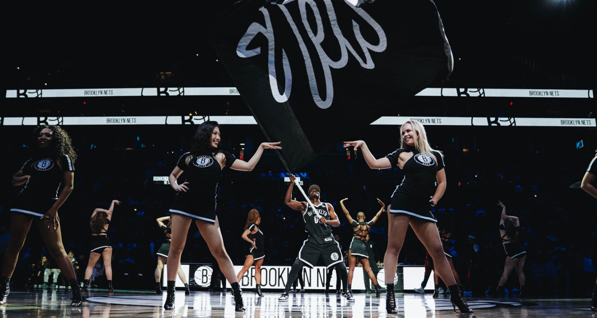On a darkly lit basketball court, members of a dance team stand proudly across the court in sassy poses as a man waves a large Nets fan in the middle.