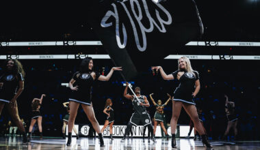 On a darkly lit basketball court, members of a dance team stand proudly across the court in sassy poses as a man waves a large Nets fan in the middle.