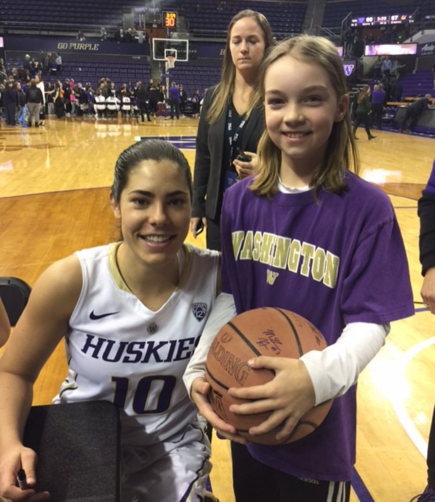 University of Washington guard Kelsey Plum squats down to take a photo with young fan Sarah Lessig. Lessig wears a purple Washington T-shirt and holds a basketball in two hands.