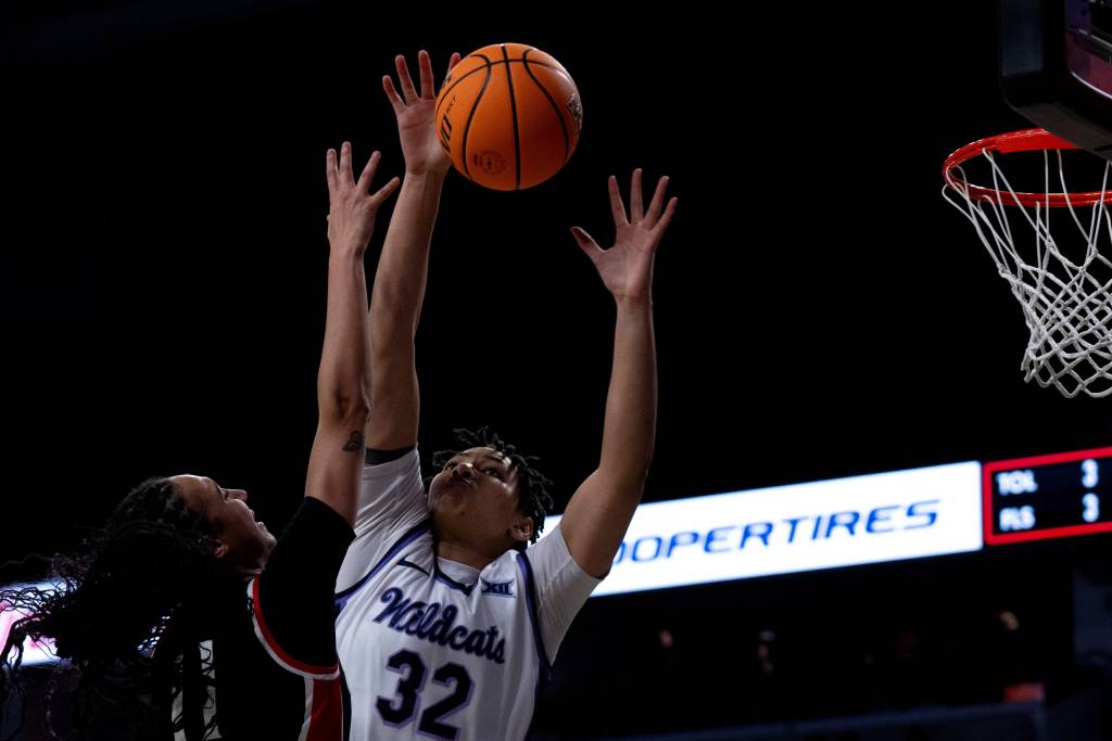 Basketball player Imani Lester blocking a shot from an opposing player. Lester wearing a white and purple basketball jersey that says "Wildcats" with the number 32. Both hands up in the air blocking a layup attempt at the basket. 