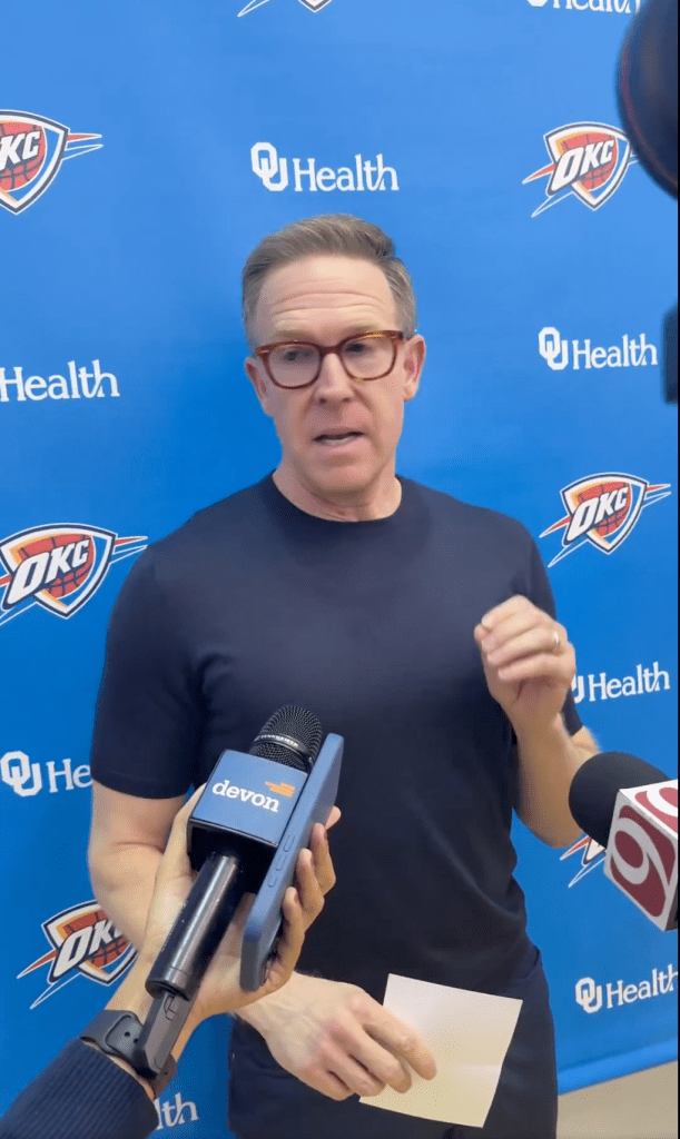 A man with glasses speaks into a "Devon" microphone during a press conference with OKC and OU Health logos in the background.