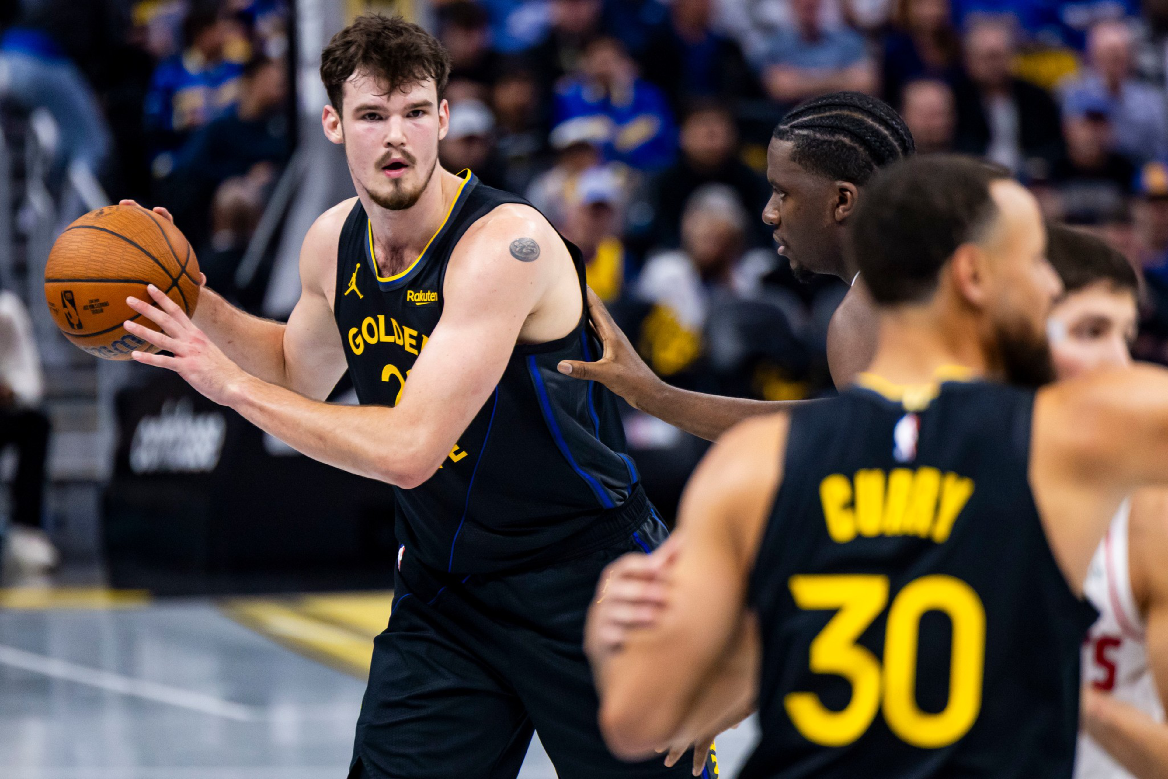 A Golden State Warriors player holds a basketball, guarded closely by an opponent, while teammate Stephen Curry, wearing number 30, is nearby.