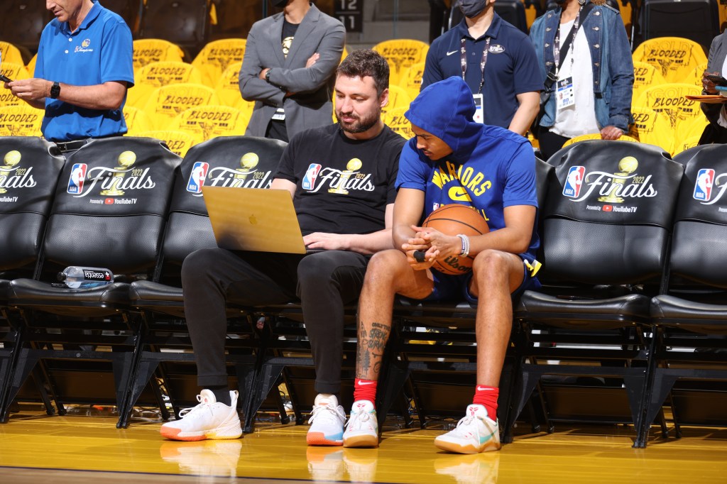 A man in a black shirt looks at a laptop while a basketball player in a blue hoodie holds a basketball and looks down, both seated on the sidelines during the 2022 NBA Finals.