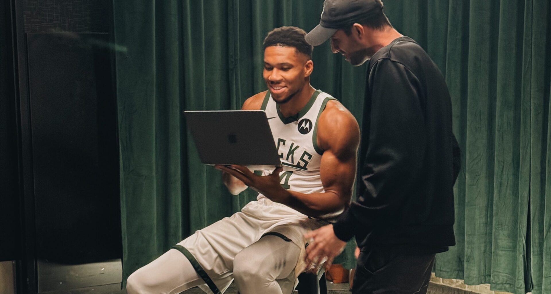 A basketball player in uniform sits on a stool holding a laptop, while another person stands next to him. Both appear to be looking at the laptop screen.