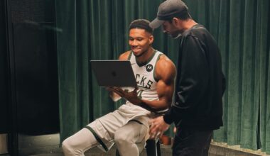 A basketball player in uniform sits on a stool holding a laptop, while another person stands next to him. Both appear to be looking at the laptop screen.