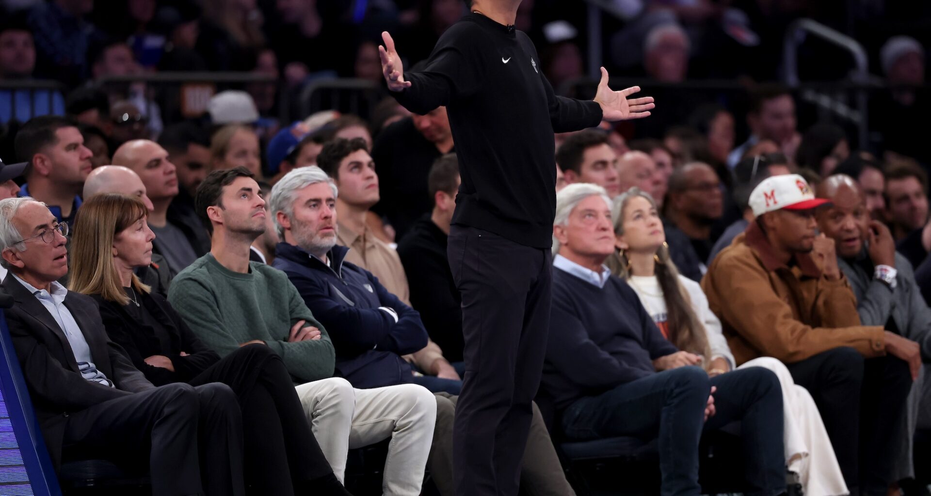 An NBA basketball coach dressed all in black stands on the sideline with his arms outstreched as if in supplication