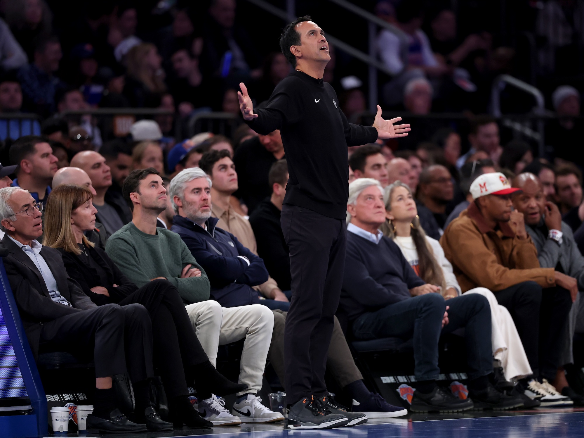 An NBA basketball coach dressed all in black stands on the sideline with his arms outstreched as if in supplication
