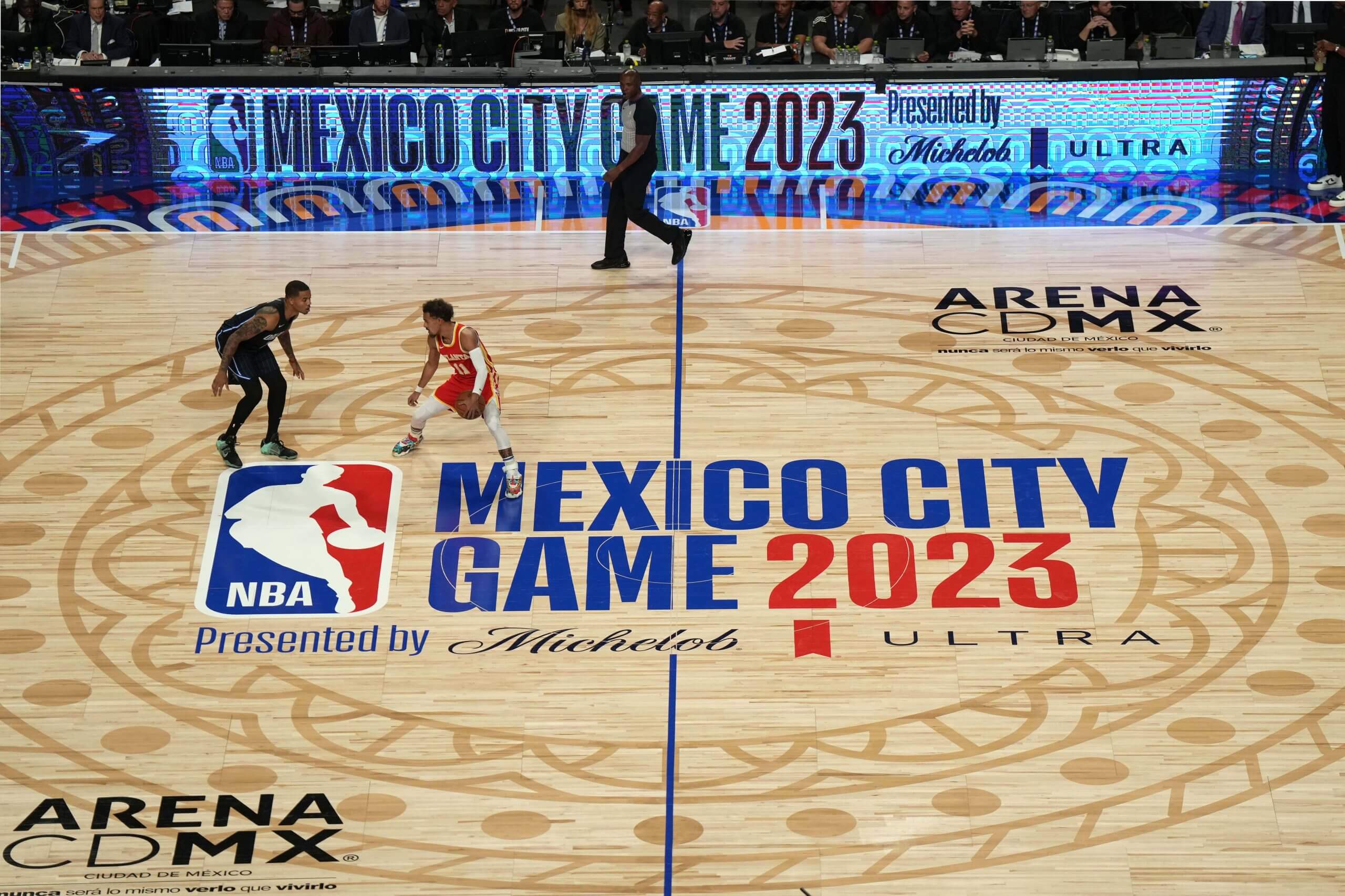 A wide shot at center court during the NBA's Mexico City game. Trae Young, dribbling the ball in a red jersey, is guarded by Markelle Fultz, who's in a black uniform.