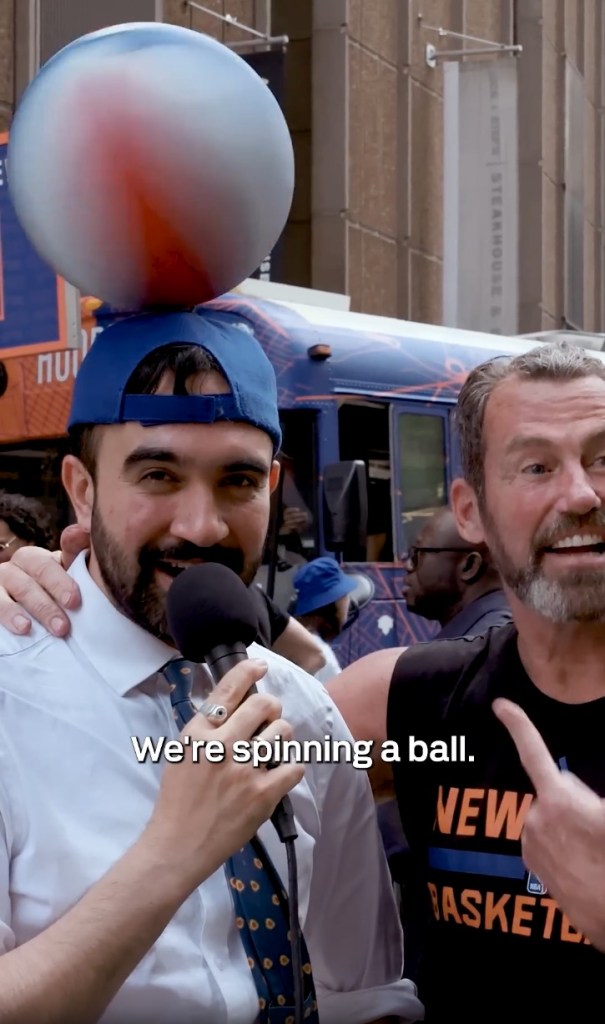 Zohran Mamdani in a backwards blue cap with a blue, white, and orange ball spinning on it, holding a microphone while a man in a black shirt points to a New York Basketball shirt.