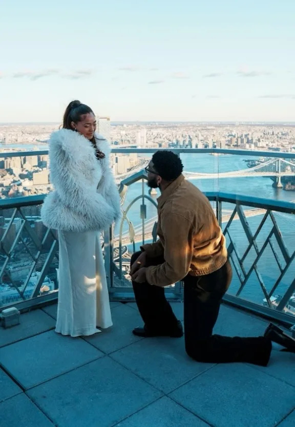 Jordyn Woods wearing a white textured coat while reacting to her engagement to Karl-Anthony Towns on a New York City rooftop