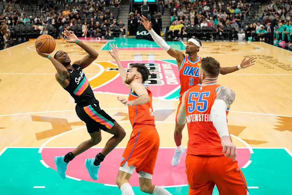 Dec 23, 2025; San Antonio, Texas, USA; San Antonio Spurs guard De’Aaron Fox (4) shoots while defended by Oklahoma City Thunder center Luke Kornet (7) and guard Shai Gilgeous-Alexander (2) during the second half at Frost Bank Center. Mandatory Credit: Scott Wachter-Imagn Images