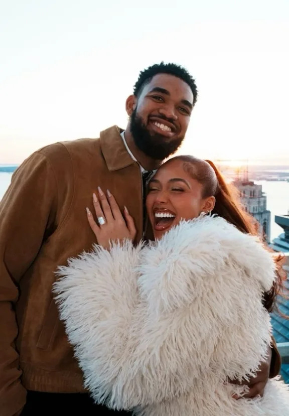 Jordyn Woods and Karl-Anthony Towns smiling together on a New York City rooftop as Woods shows her engagement ring