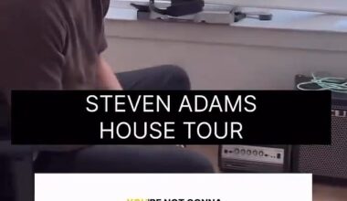 Adams is seen sitting in a small-yet-sparsely-furnished room as he gestures to the floor