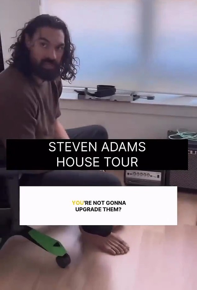 Adams is seen sitting in a small-yet-sparsely-furnished room as he gestures to the floor