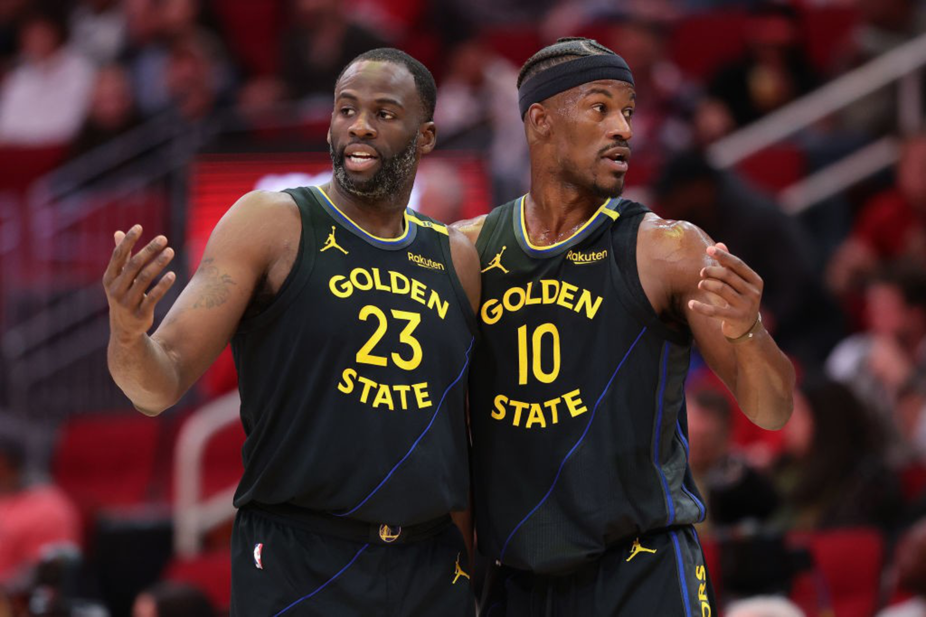 Two basketball players in dark jerseys with “Golden State” are on the court. One wears number 23, and the other 10, both gesturing and focused on the game.