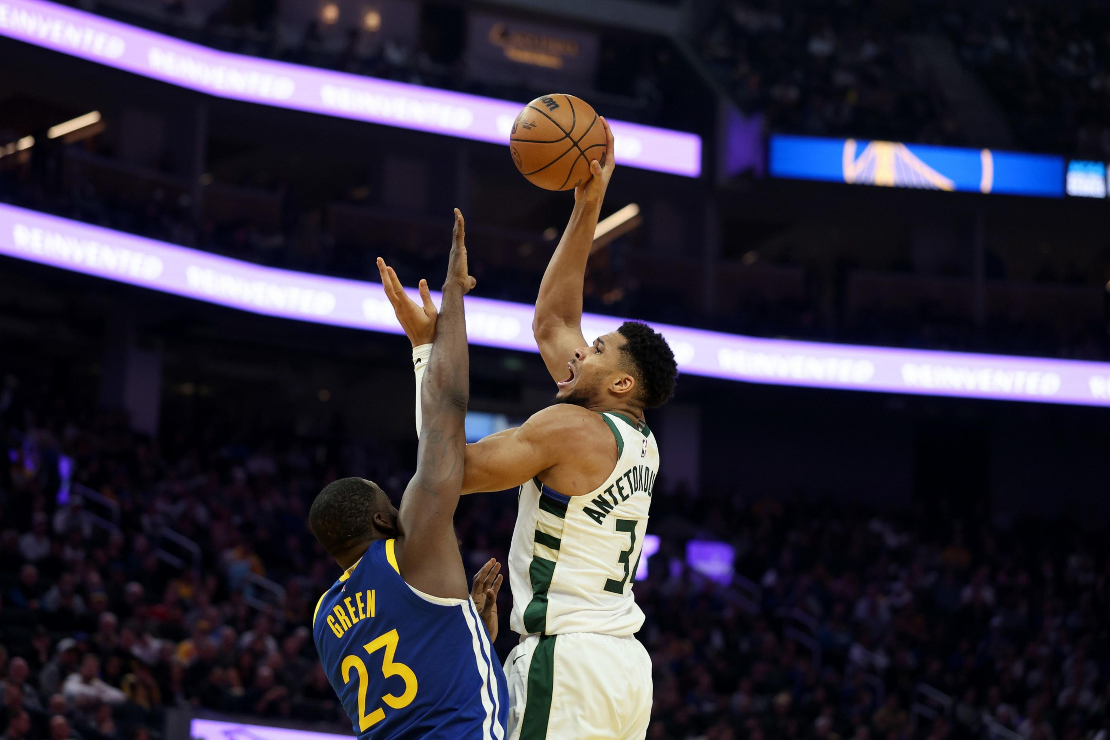 Giannis Antetokounmpo jumps to shoot a basketball over Draymond Green, who is reaching up to block the shot during a game.