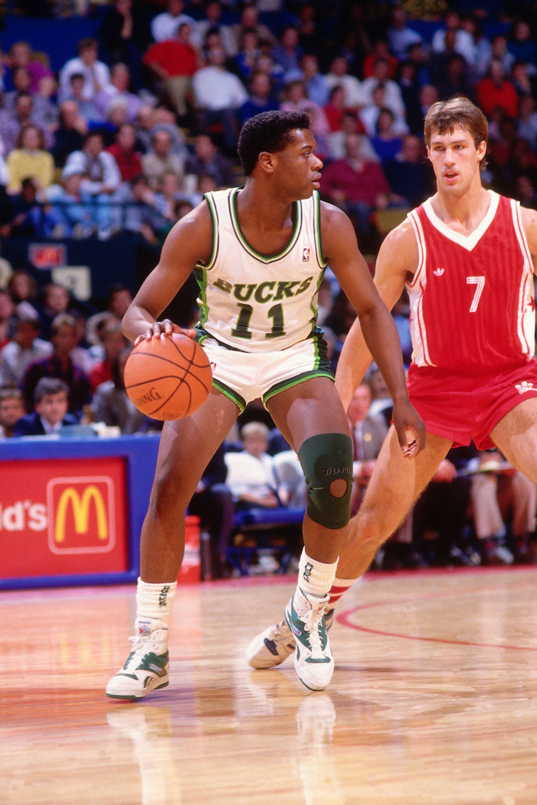 A basketball player in a green and white Bucks uniform dribbles the ball while being guarded by an opponent in a red uniform during a game.