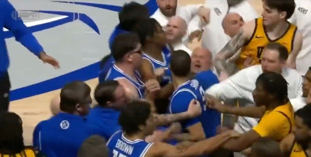 A basketball player in a blue jersey is held back from an opposing player in a yellow jersey, while coaches and other players attempt to de-escalate the situation.
