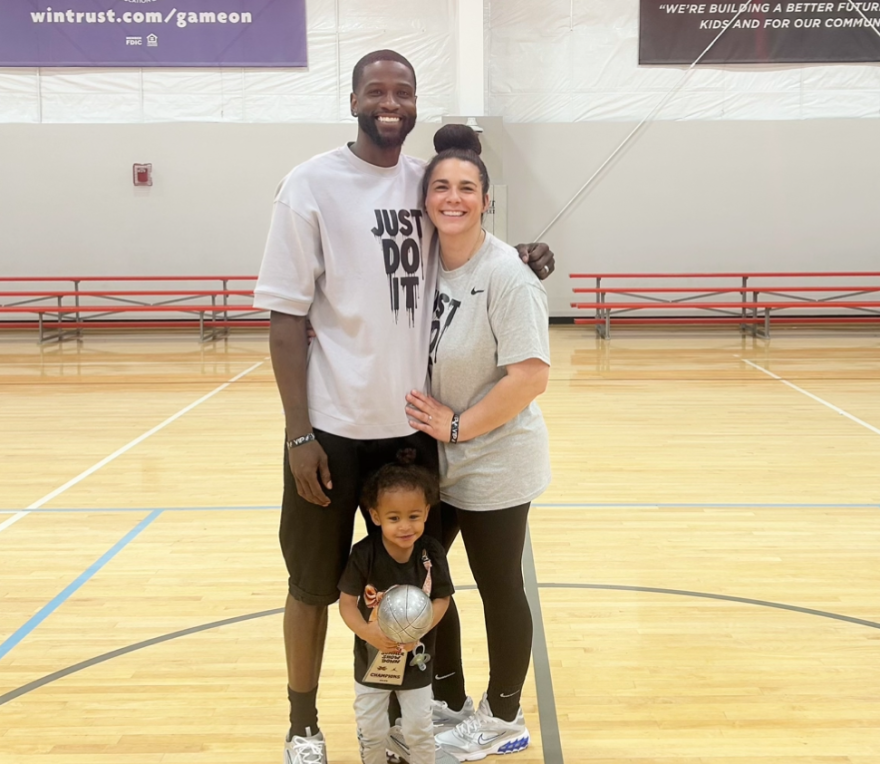 Man and woman posing for a photo with a young child standing between them and holding a basketball trophy in a gymnasium