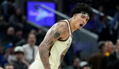 A basketball player with tattoos and a headband is shouting passionately during a game, with a crowd blurred in the background.