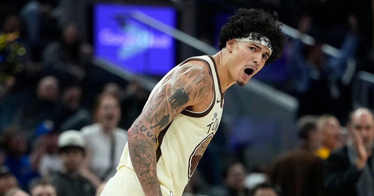 A basketball player with tattoos and a headband is shouting passionately during a game, with a crowd blurred in the background.